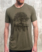 Man wearing olive Tree and Crumbling City graphic tee with tree roots over cityscape illustration