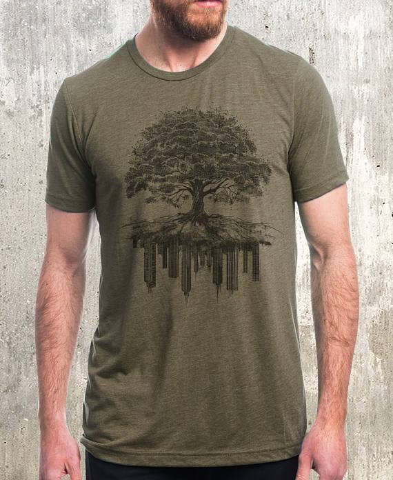 Man wearing olive Tree and Crumbling City graphic tee with tree roots over cityscape illustration