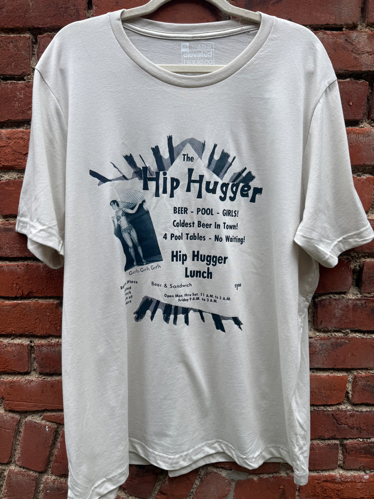 The Original Kokomo Hip Hugger Ad tee in natural, hung against a brick wall